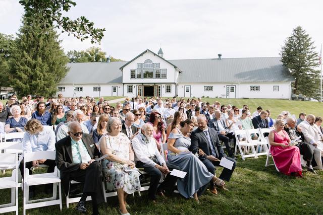 Guests seated for an outdoor ceremony on the lawn in front of the white barn
