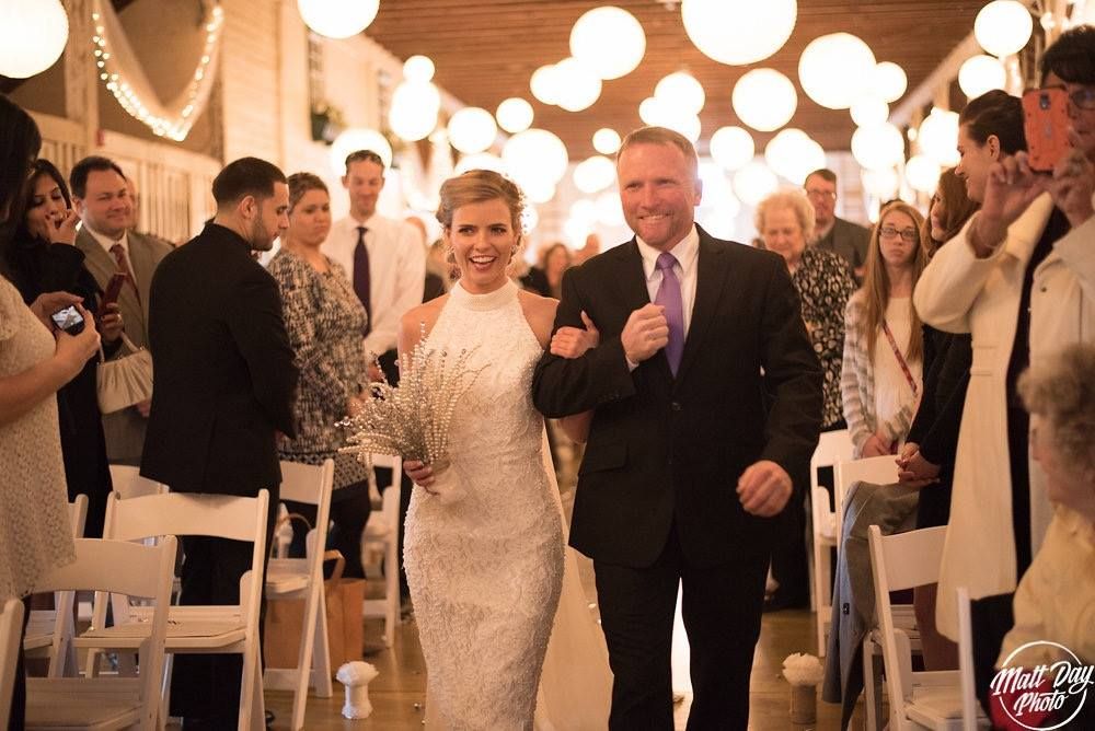 Father walks the bride down the aisle inside the barn as guests look on