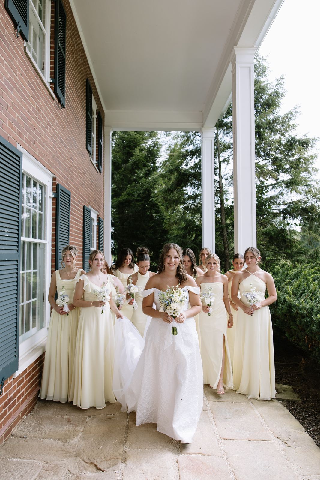 Bride walking with her bridesmaids along the porch of the brick house