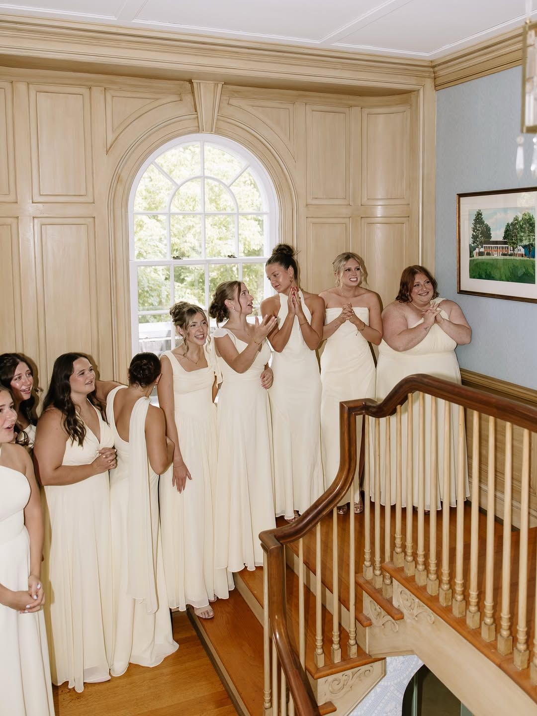 Bridesmaids in ivory gowns react with joy on the grand staircase of the house