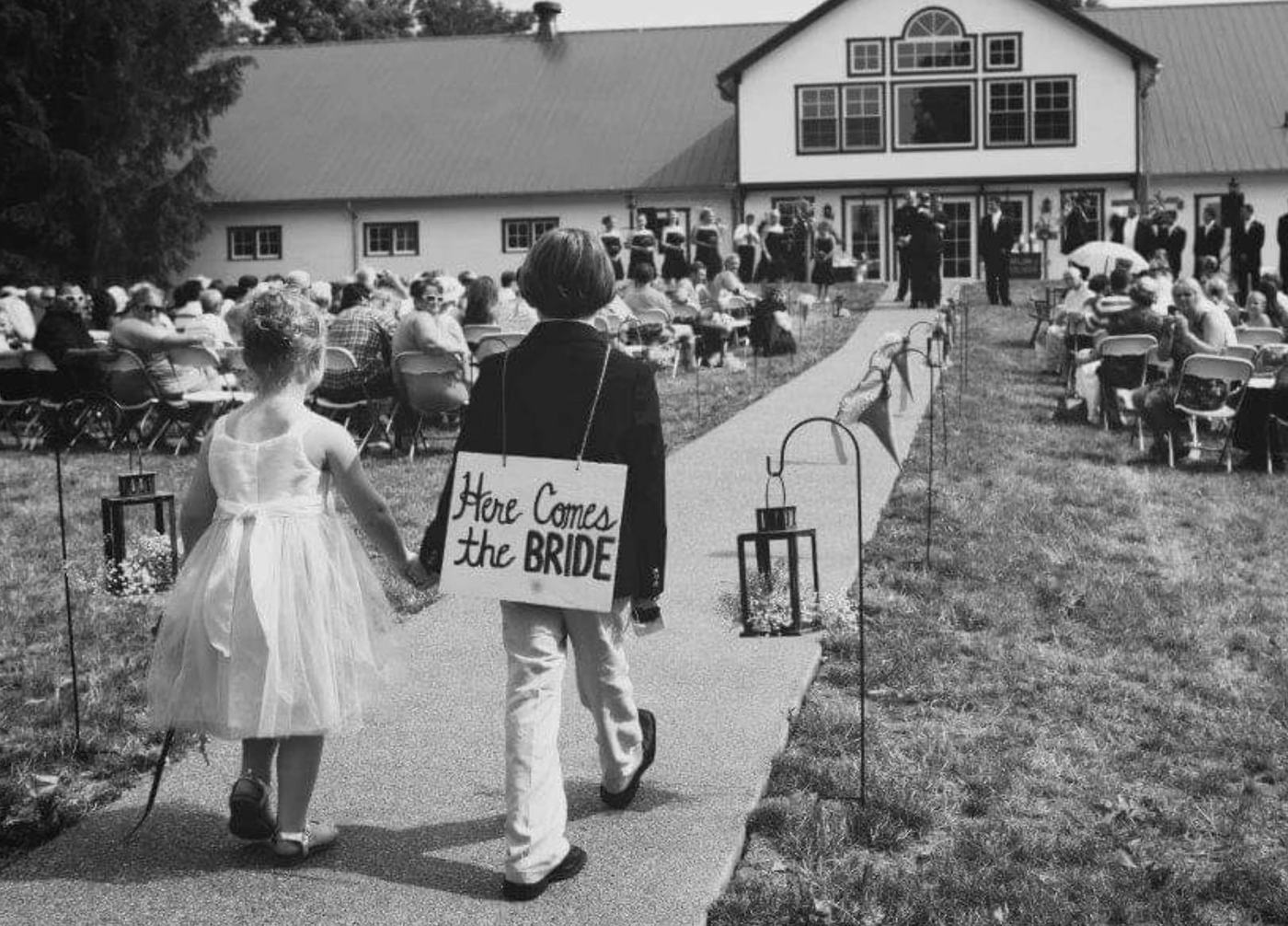 Ring bearer with a Here Comes the Bride sign and flower girl walking the aisle toward the barn